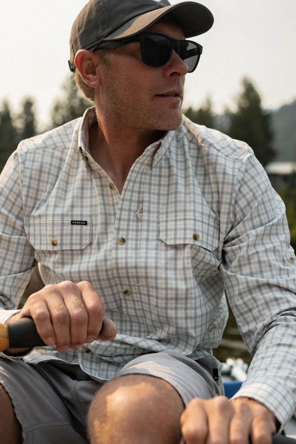 A man wearing a light beige and white long-sleeve plaid button-down shirt, gray shorts, a dark cap, and sunglasses, holding a paddle in an outdoor setting.