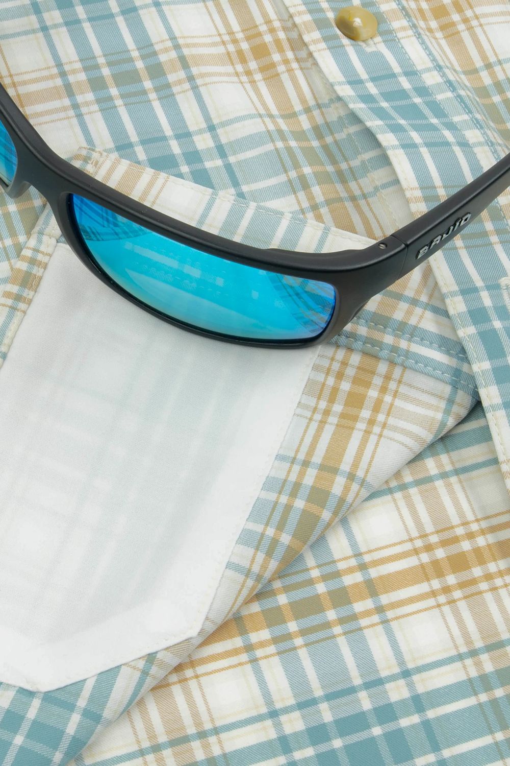 Close-up of black sunglasses with blue mirrored lenses resting on a light blue, beige, and white plaid button-down shirt.