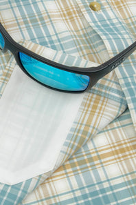 Close-up of black sunglasses with blue mirrored lenses resting on a light blue, beige, and white plaid button-down shirt.