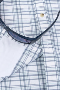 A close-up of a white plaid button-down shirt with blue-gray stripes, featuring a folded white pocket square and a pair of dark sunglasses resting on top.