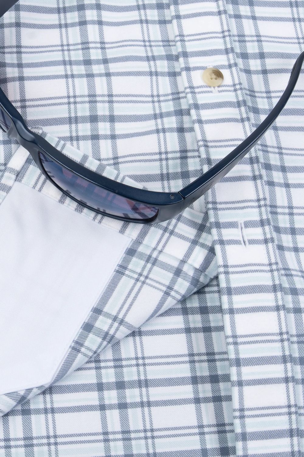 A close-up of a white plaid button-down shirt with blue-gray stripes, featuring a folded white pocket square and a pair of dark sunglasses resting on top.