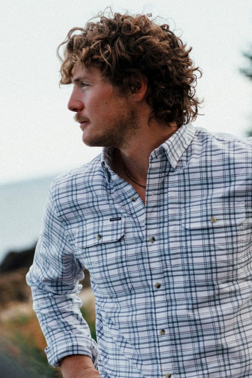 A man with curly brown hair wearing a blue and white plaid button-down shirt, photographed outdoors with a soft-focus natural background.