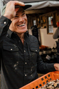 A smiling man wearing a dark button-up shirt and cap, holding an orange crate in a rustic indoor setting.