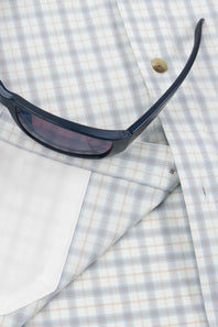 A close-up of a light blue and white plaid button-down shirt with a white pocket square and a pair of dark sunglasses resting on top.