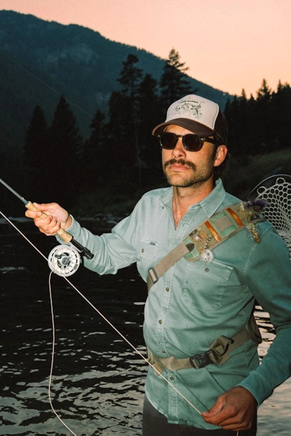 A man wearing a light blue button-down shirt, sunglasses, and a baseball cap stands in a river holding a fly fishing rod, with mountains and trees in the background at sunset.