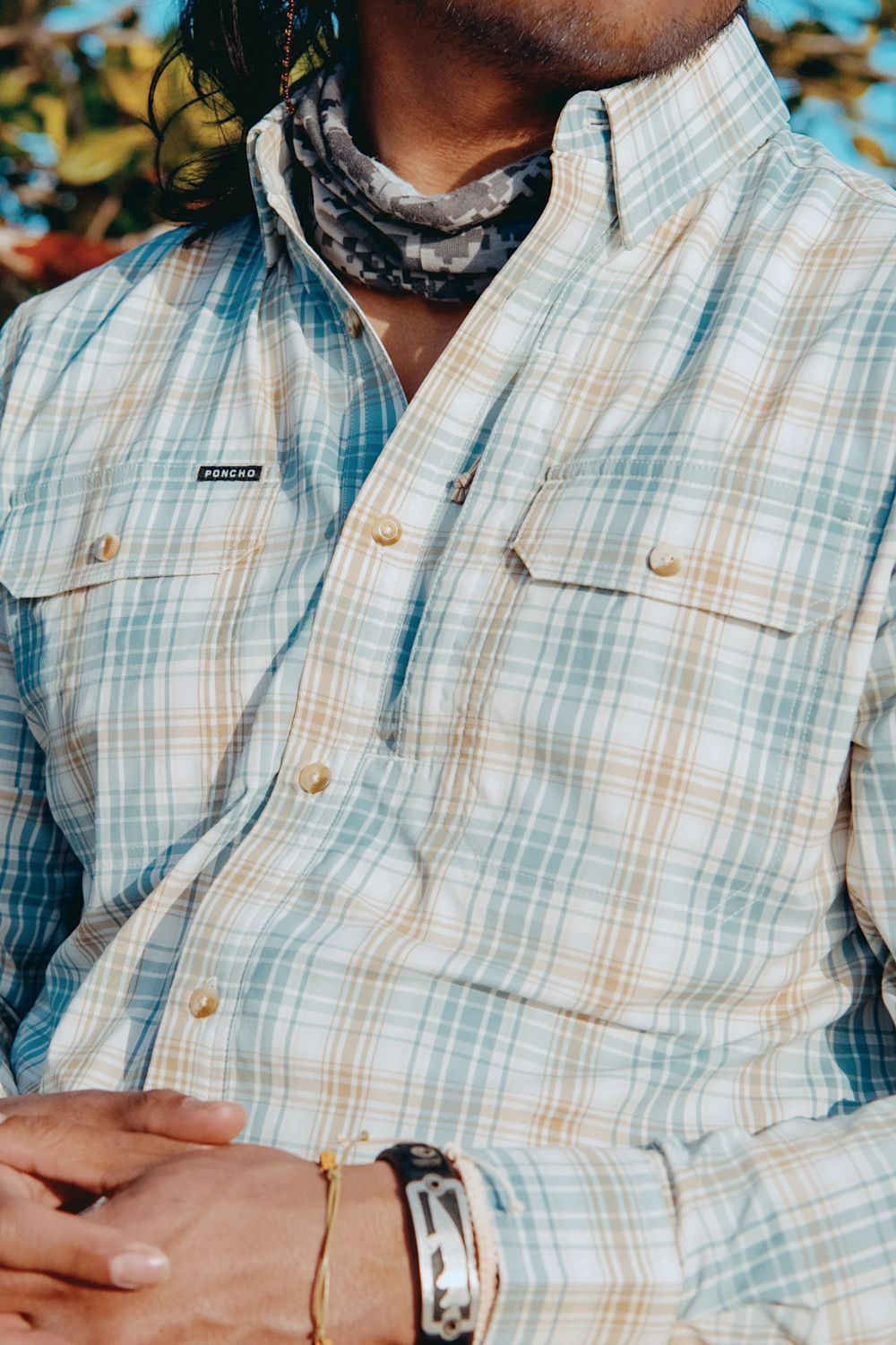 A close-up of a person wearing a light blue and beige plaid button-up shirt with a gray patterned neck gaiter. The shirt has two chest pockets with button flaps and a small black brand patch on the left chest pocket.