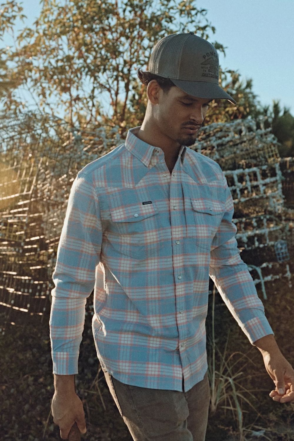 A man wearing a light blue and white plaid long-sleeve button-down shirt, brown pants, and a gray baseball cap, standing outdoors in front of a rustic wooden fence.
