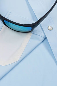 Close-up of light blue dress shirt fabric with a white pocket and a pair of black sunglasses resting across the chest.