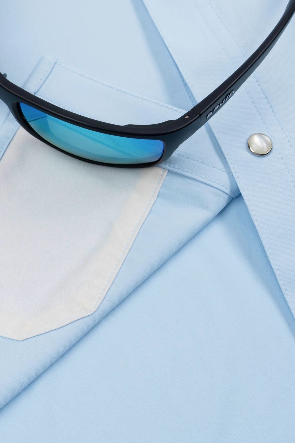 Close-up of light blue dress shirt fabric with a white pocket and a pair of black sunglasses resting across the chest.
