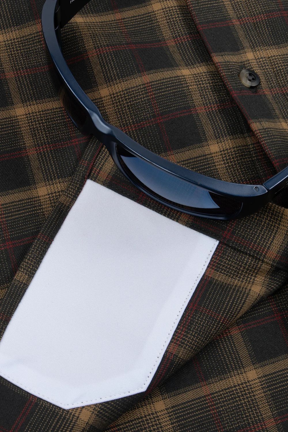 A close-up flat lay of a plaid shirt with a white dress shirt cuff and a pair of dark sunglasses resting on top.