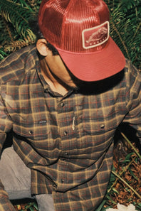 A man wearing a red trucker hat with a rectangular patch and a brown and red plaid button-up shirt, photographed outdoors among green foliage.