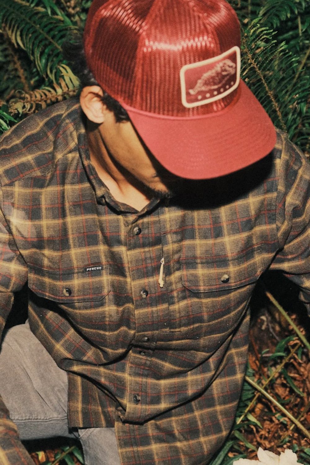 A man wearing a red trucker hat with a rectangular patch and a brown and red plaid button-up shirt, photographed outdoors among green foliage.