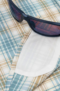 A close-up of a plaid button-down shirt in light blue, beige, and white, with a white pocket square tucked into the collar and dark sunglasses resting on top.