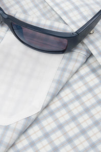 A close-up of a light blue and white plaid button-down shirt with a white pocket, styled with a pair of black sunglasses on top.