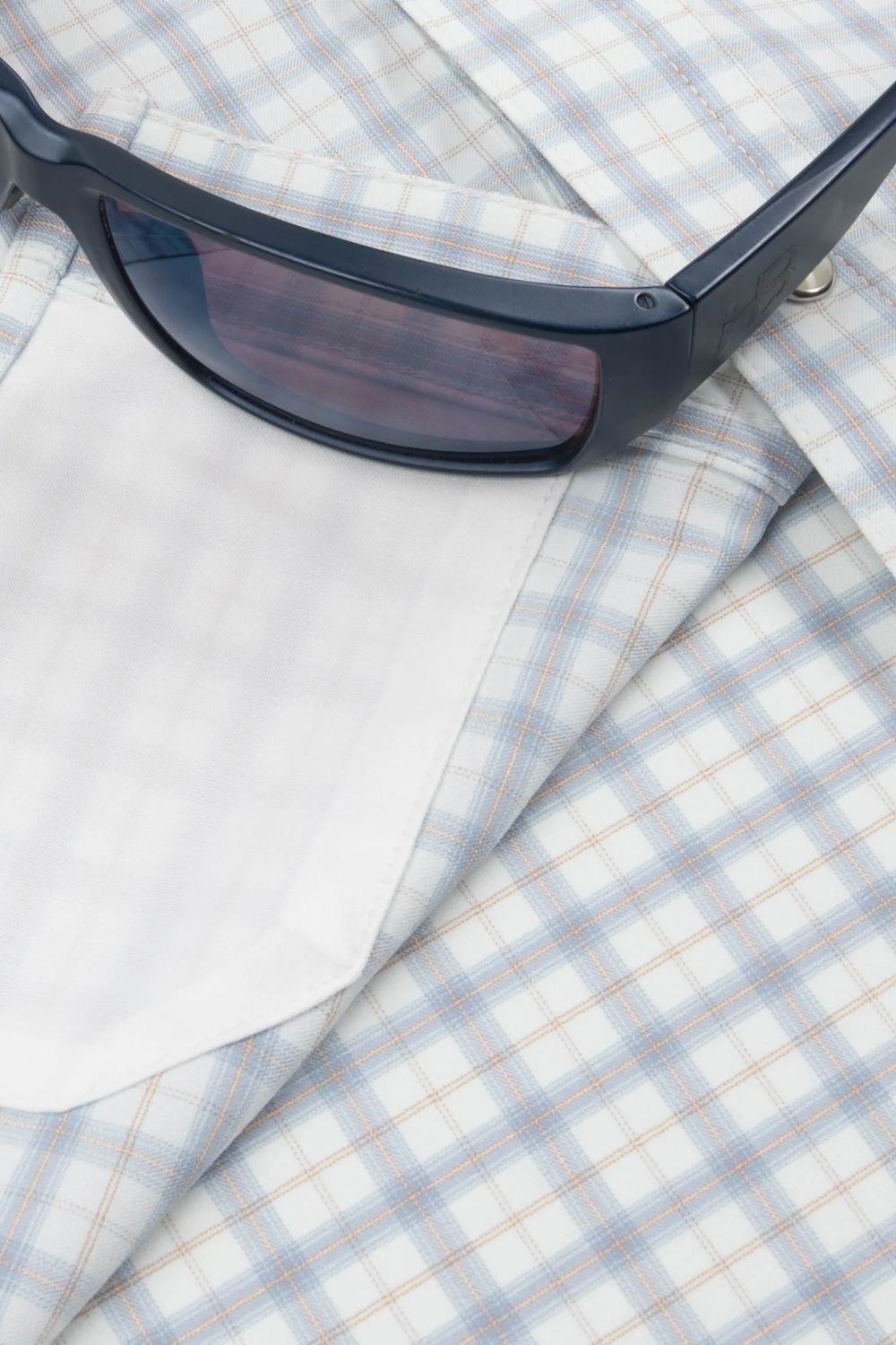 A close-up of a light blue and white plaid button-down shirt with a white pocket, styled with a pair of black sunglasses on top.
