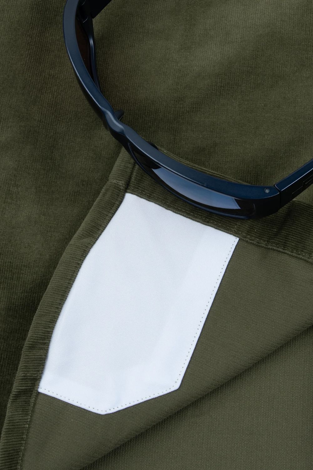 Close-up of an olive green corduroy garment with a white fabric pocket and a pair of dark sunglasses resting on top.