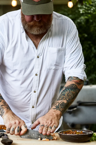 A bearded man wearing a white button-down shirt and a green cap is shown from the chest up, actively slicing a fish on a wooden cutting board with a large knife. A small bowl of food is visible in the foreground, with a blurred outdoor background of green foliage and warm hanging lights.