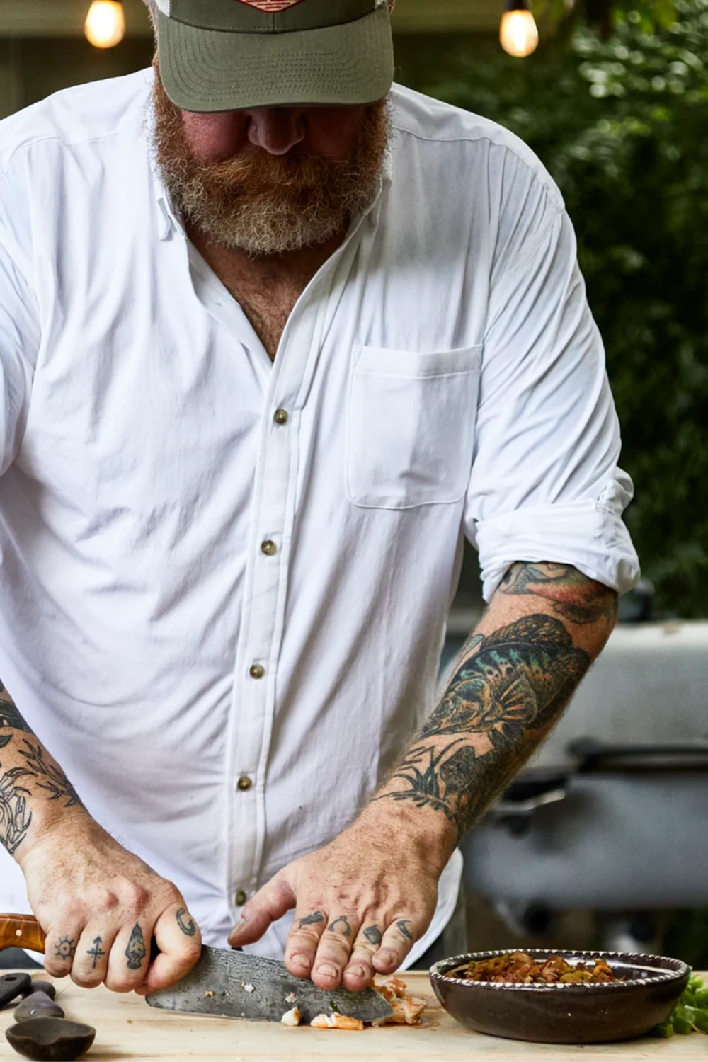 A bearded man wearing a white button-down shirt and a green cap is shown from the chest up, actively slicing a fish on a wooden cutting board with a large knife. A small bowl of food is visible in the foreground, with a blurred outdoor background of green foliage and warm hanging lights.