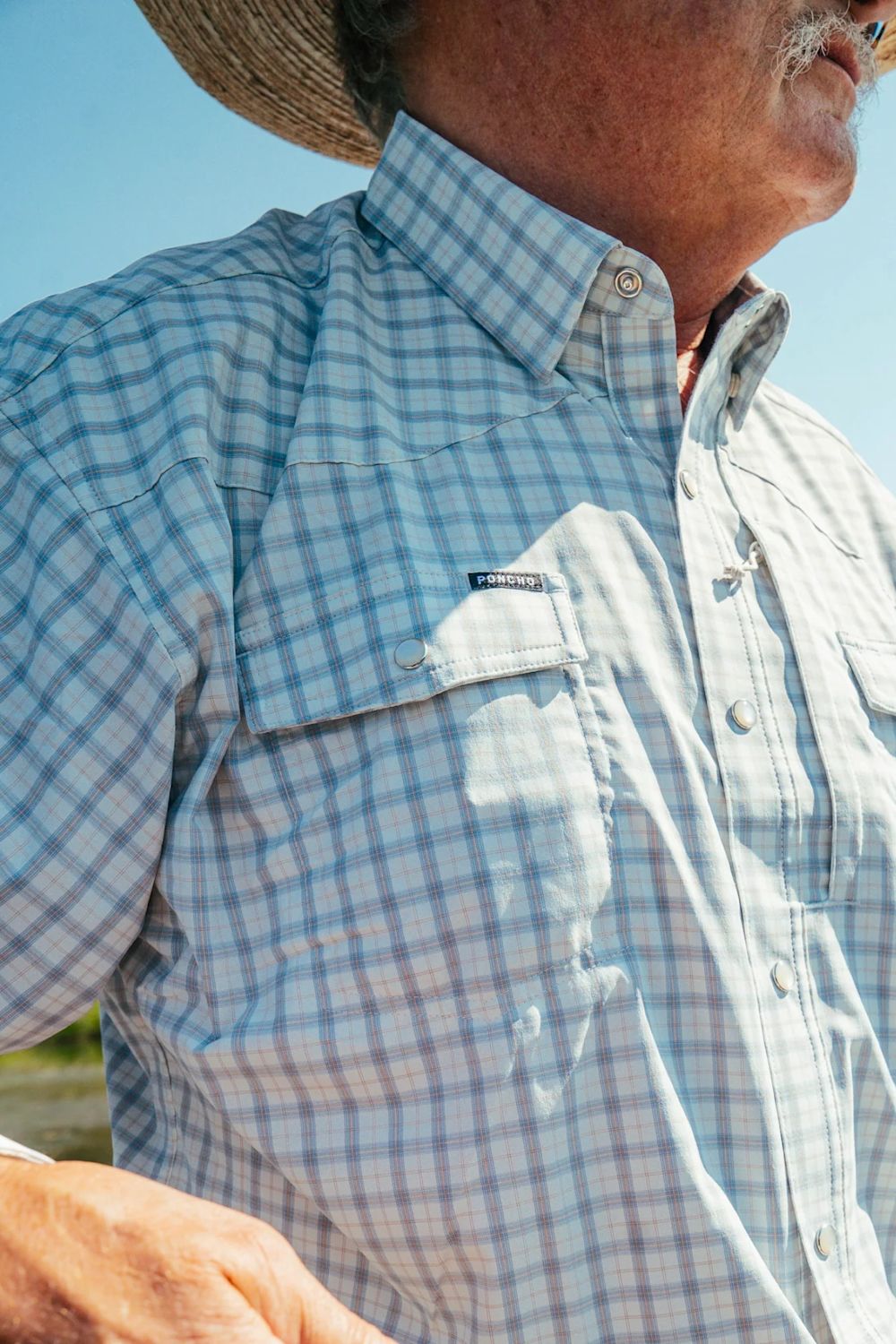 A close-up of a man wearing a light blue and white plaid button-down shirt with a chest pocket, under bright outdoor sunlight.