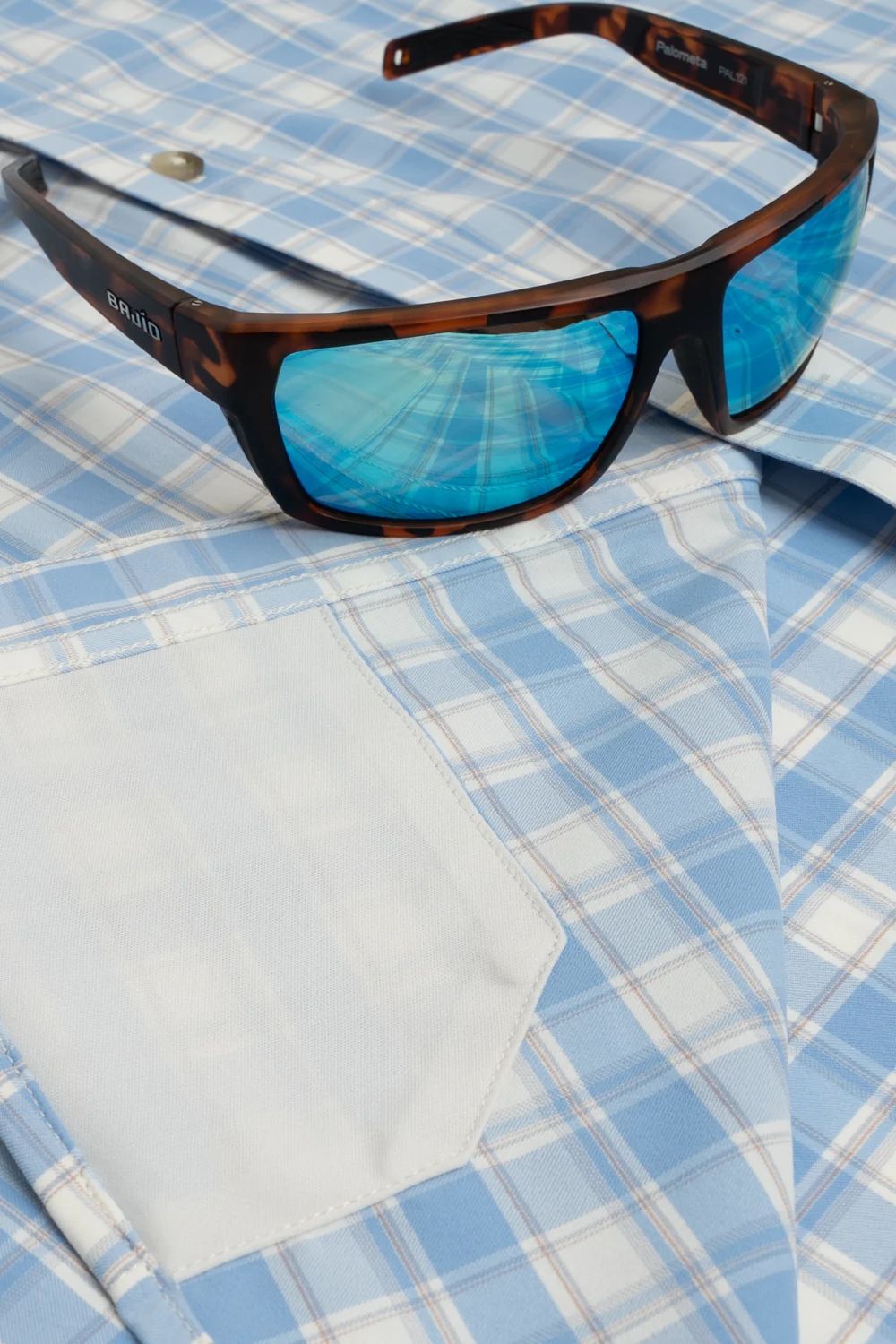 Tortoiseshell sunglasses with blue mirrored lenses resting on a light blue and white plaid button-down shirt.