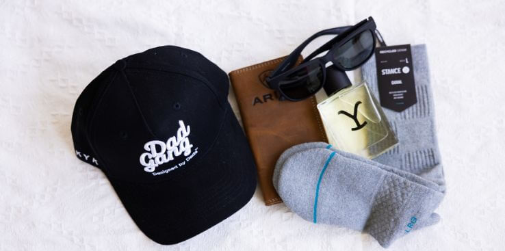 Black cap with 'Dad Gang' text, wallet sunglasses, cologne and socks on a white surface