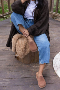 A person sitting on a wooden stool on a wooden deck, wearing a dark brown knit cardigan, white t-shirt, blue jeans, and tan slip-on mule shoes. A brown faux-fur accessory rests on the stool.