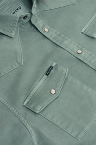 Close-up of a sage green western-style shirt jacket with silver snap buttons and a chest pocket, showing the brand label 'Poncho' on the collar 