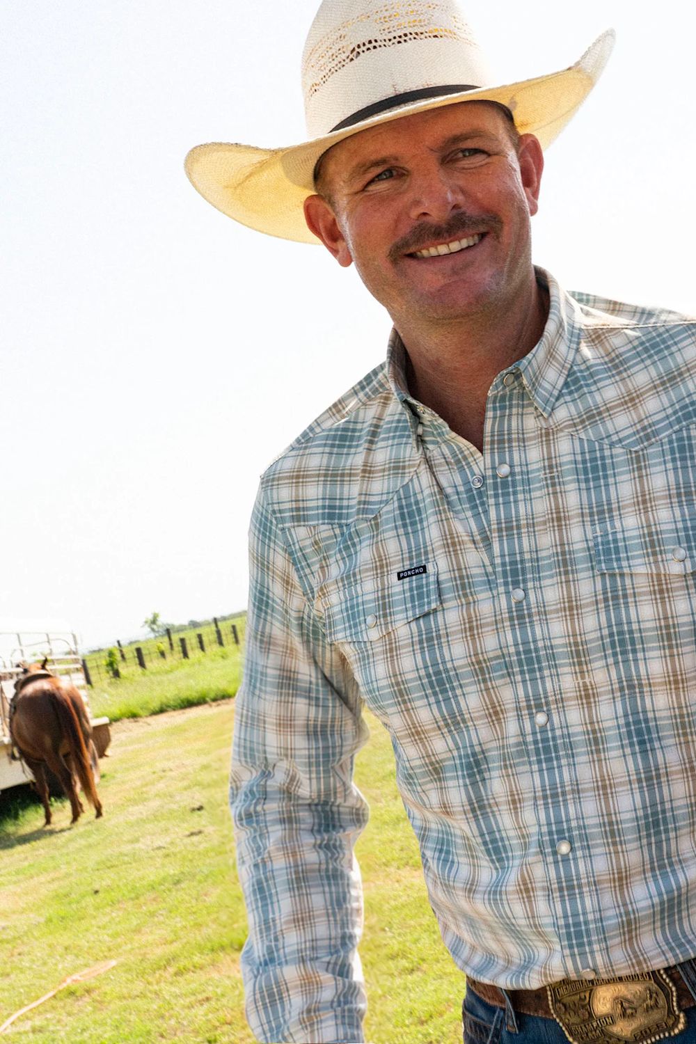 A smiling man wearing a cream cowboy hat and a blue, white, and tan plaid long-sleeve button-down shirt, standing outdoors in a grassy field with a cow in the background.