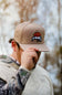 A man wearing a tan baseball cap with a dog patch on the front, holding the brim with one hand. The background is a soft-focus outdoor scene with trees.