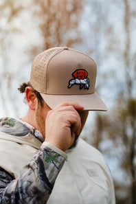 A man wearing a tan baseball cap with a dog patch on the front, holding the brim with one hand. The background is a soft-focus outdoor scene with trees.