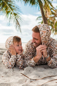 YOUTH-BURLEBO-PERFORMANCE-HOODIE-VENADO-CAMO-Y-PH-VENC-BROWN_3 Father and son lying on a beach wearing matching camouflage hoodies with palm trees in the background.