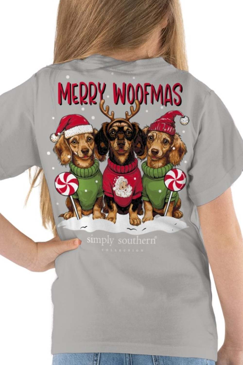 A woman wearing a light gray t-shirt, viewed from the back. The shirt features a large graphic of three dachshunds dressed in Christmas sweaters and Santa hats, with the text 'MERRY WOOFMAS' and the brand 'simply southern' printed below.