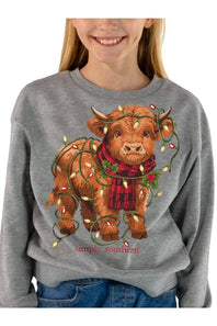 A woman wearing a heather gray crewneck sweatshirt with a graphic of a Highland cow wearing a red plaid scarf and holiday lights, with the text 'simply southern' below it.