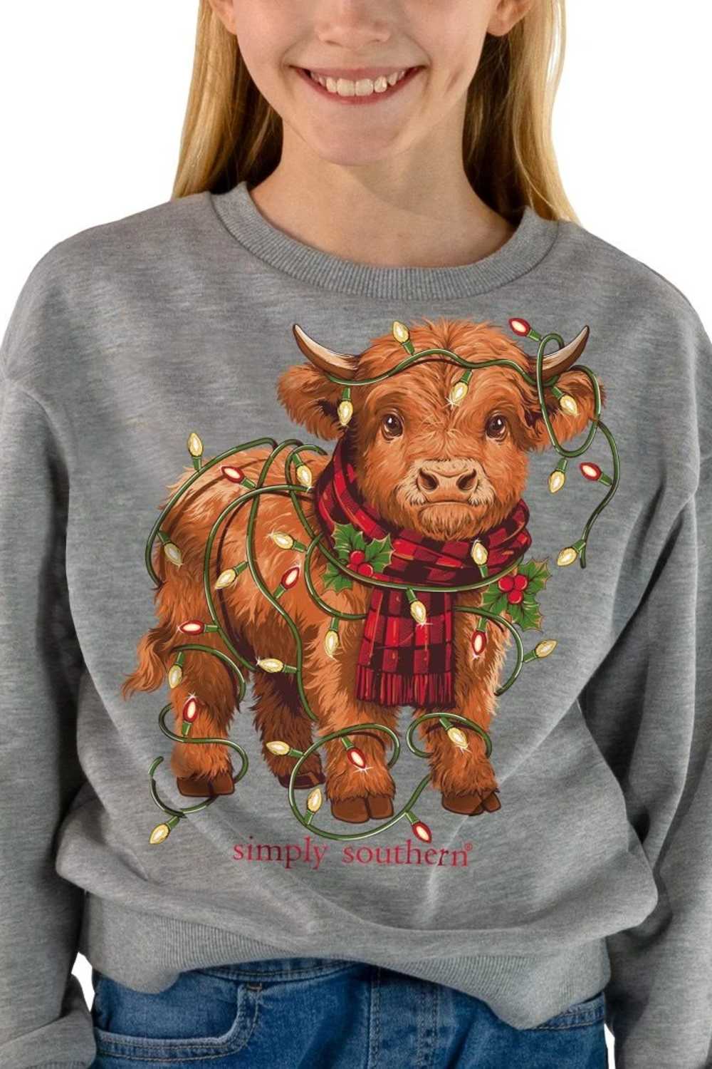 A person wearing a heather gray crewneck sweatshirt featuring a graphic of a brown Scottish Highland cow decorated with Christmas lights and a red plaid scarf, with the text 'simply southern' below.