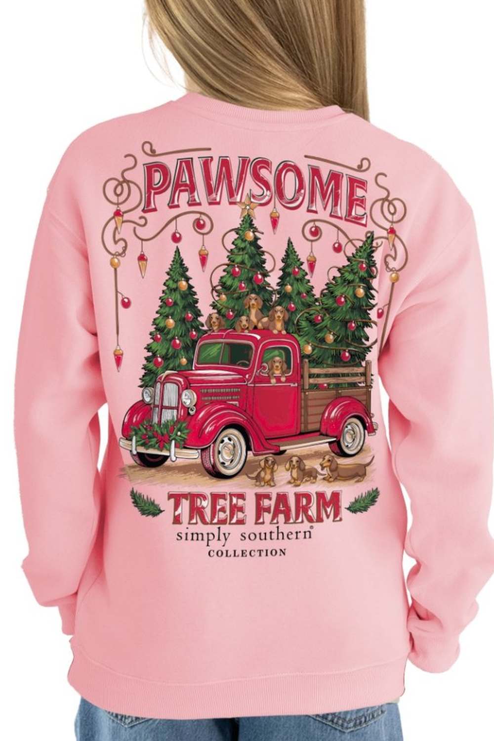 A person wearing a light pink crewneck sweatshirt, shown from the back. The sweatshirt features a large graphic print of a red vintage truck carrying Christmas trees, with the text 'PAWSOME' and 'TREE FARM simply southern COLLECTION'.