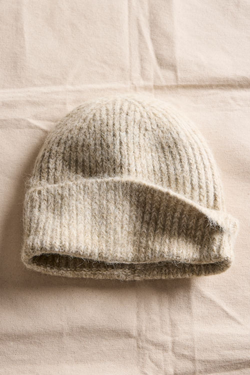 A ribbed knit beanie hat in a heathered oatmeal beige color, laid flat on a textured beige fabric background.