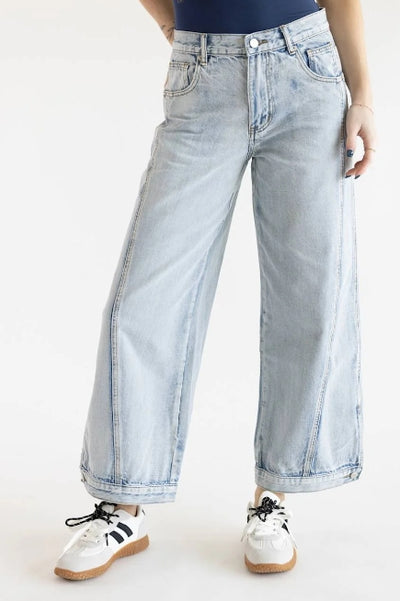 A woman wearing light wash barrel leg denim jeans with a navy top and white with black detail sneakers on a white background