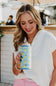 A woman in a white top and pink skirt holds a light blue can with a yellow lemon pattern and the brand name 'Katydid' printed on it, set against a blurred outdoor patio background.