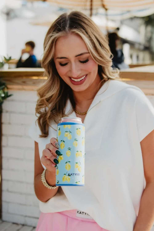 A woman in a white top and pink skirt holds a light blue can with a yellow lemon pattern and the brand name 'Katydid' printed on it, set against a blurred outdoor patio background.