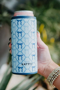 Hand holding a blue can cooler with a bow pattern and 'KATYDID' branding against a blurred natural background.