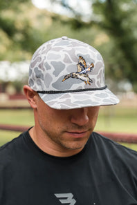 A man wearing a gray and white camouflage baseball cap with a detailed embroidered duck in flight on the front, photographed outdoors with a blurred park background.