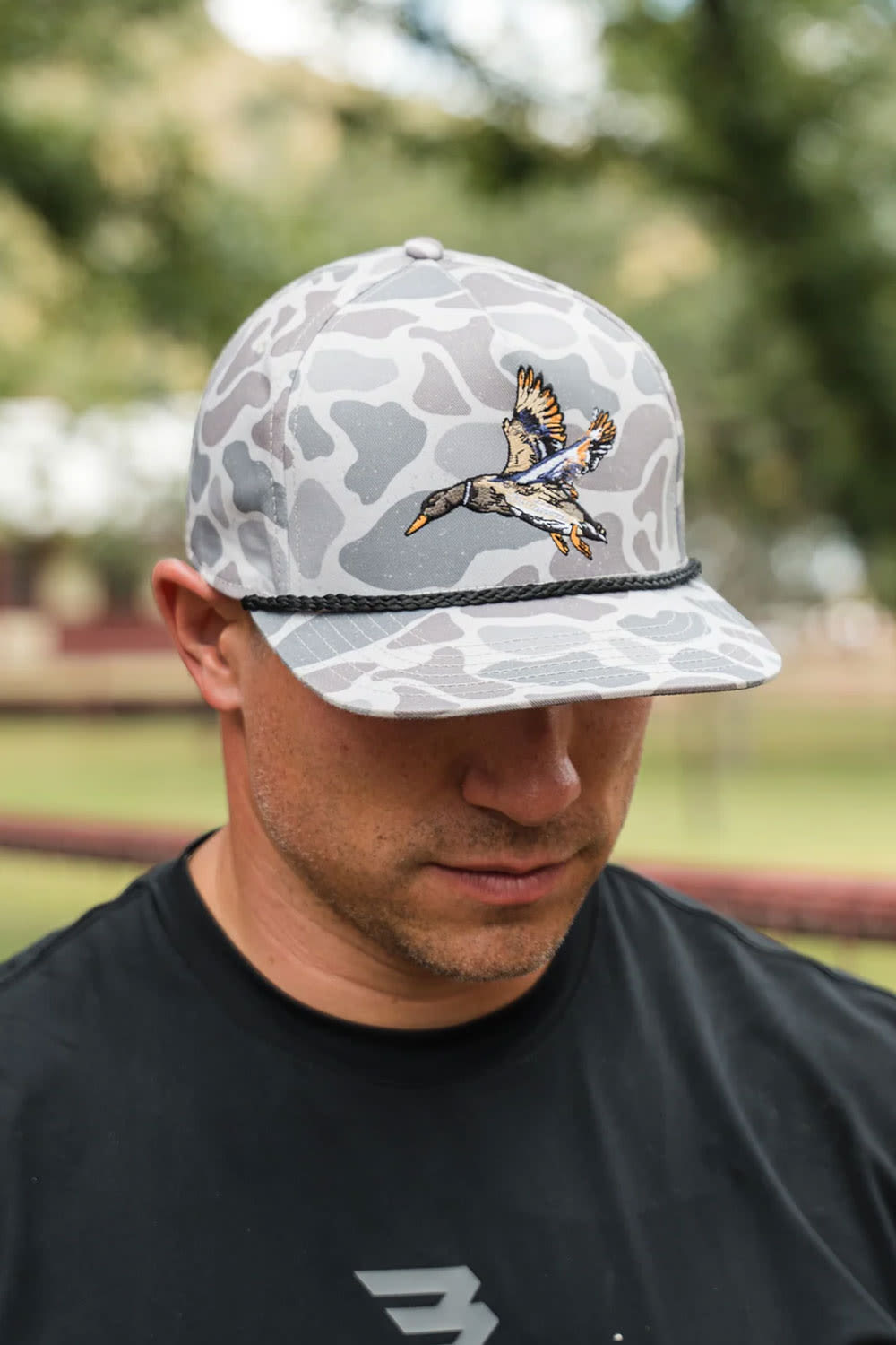 A man wearing a gray and white camouflage baseball cap with a detailed embroidered duck in flight on the front, photographed outdoors with a blurred park background.