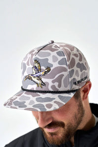 A man wearing a camouflage baseball cap with a flying duck graphic and a black rope detail under the brim. The cap features a light gray and brown camo pattern, and the brand name 'BURLEBO' is visible on the side.
