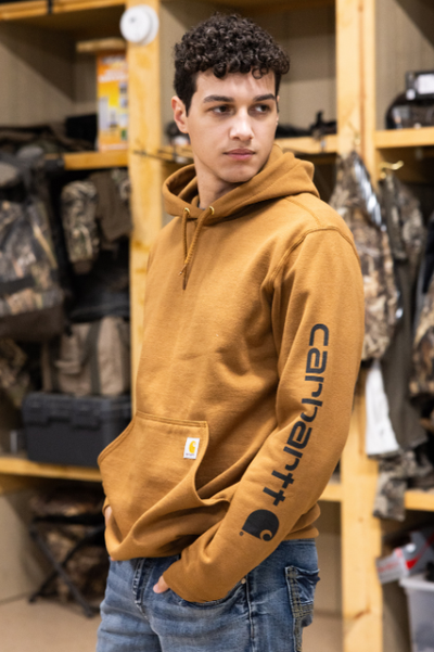 Man standing in a warehouse wearing a brown hoodie with CARHARTT down the sleeve and blue jeans