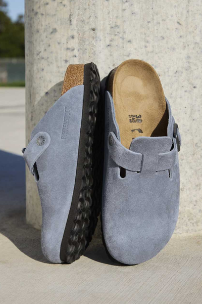 Image shows dusty blue colored clogs with black soles leaning against a beige speckled wall