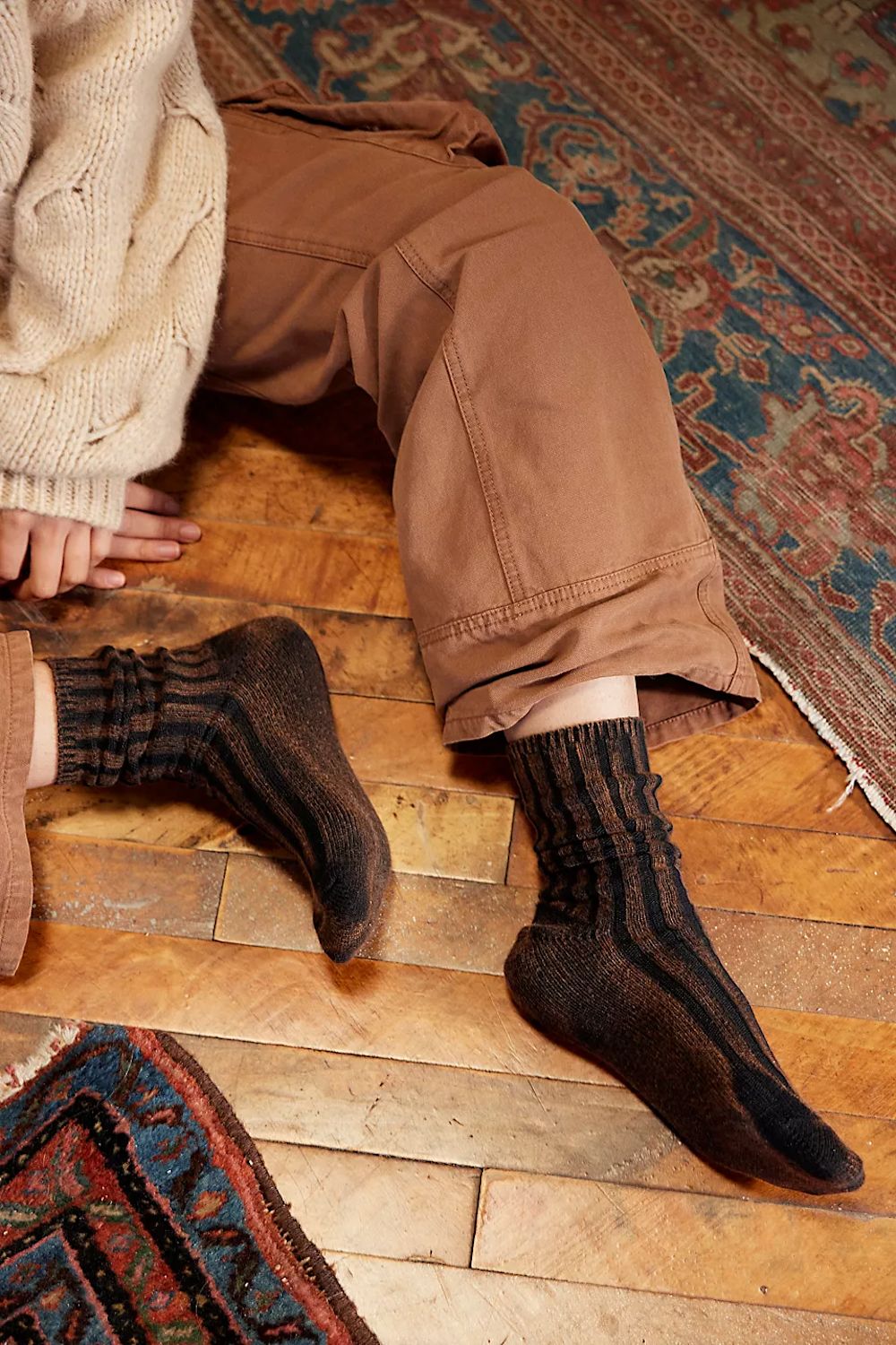 A person sitting on a wooden floor wearing dark brown and black striped ribbed socks, brown pants, and a cream cable-knit sweater, with patterned rugs visible in the background.