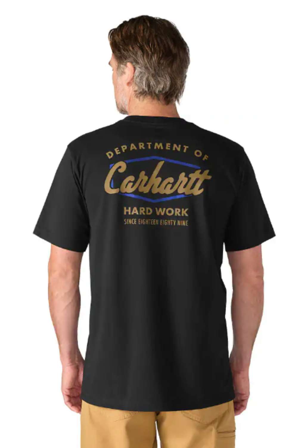 A man wearing a black Carhartt short-sleeve t-shirt with a small gold embroidered logo and the signature Carhartt square patch on the left chest, paired with tan pants, against a light gray studio background.