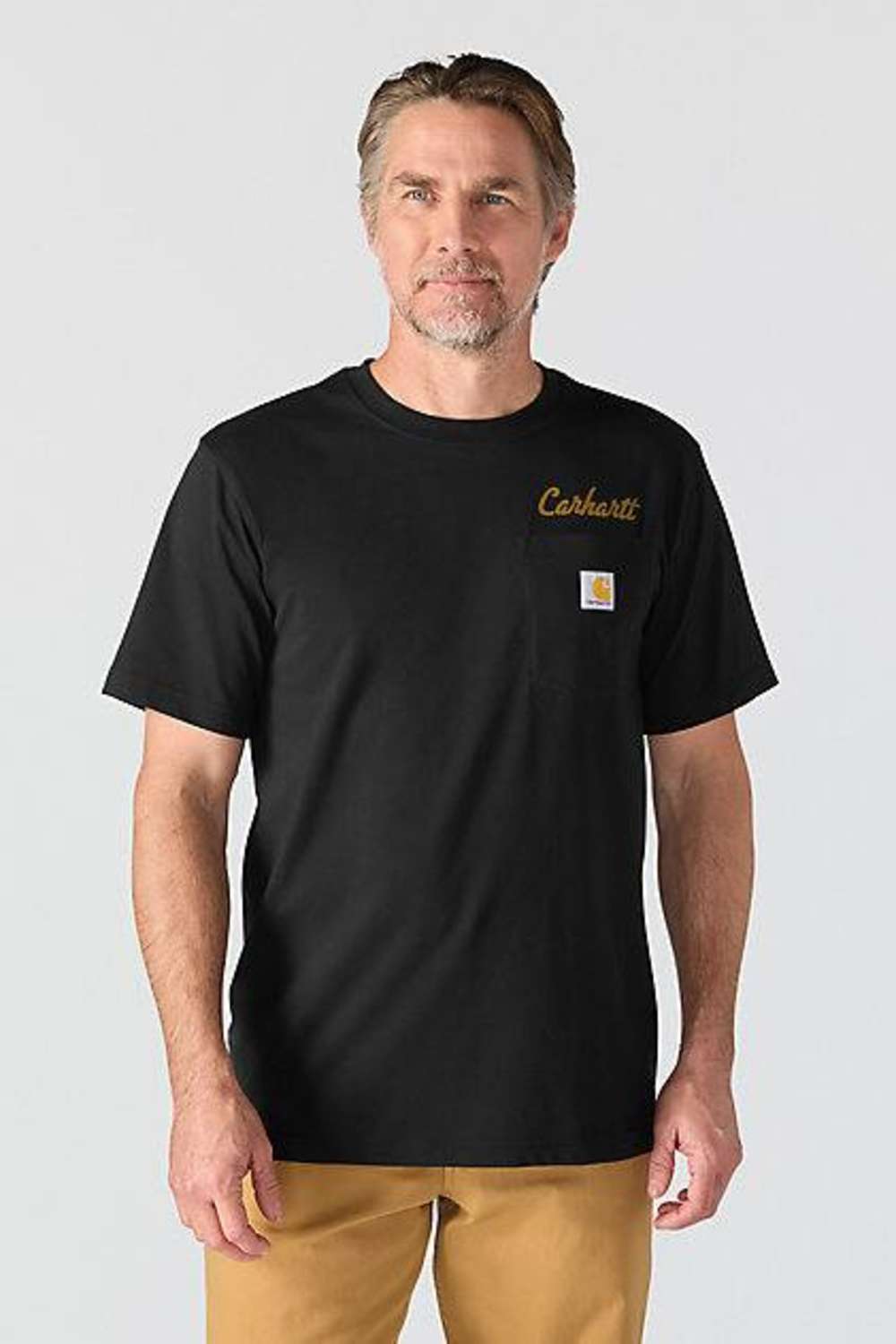 A man wearing a black Carhartt short-sleeve t-shirt with a small gold embroidered logo and the signature Carhartt square patch on the left chest, paired with tan pants, against a light gray studio background.
