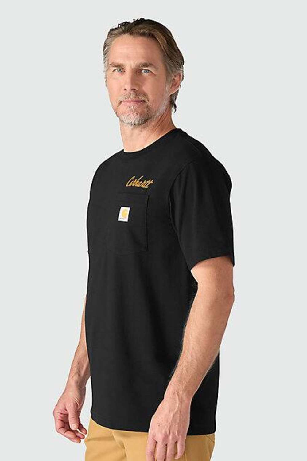 A man wearing a black Carhartt short-sleeve t-shirt with a chest pocket, featuring a gold script logo on the chest and the signature Carhartt square logo patch on the pocket. He is also wearing tan pants, set against a solid light gray background.