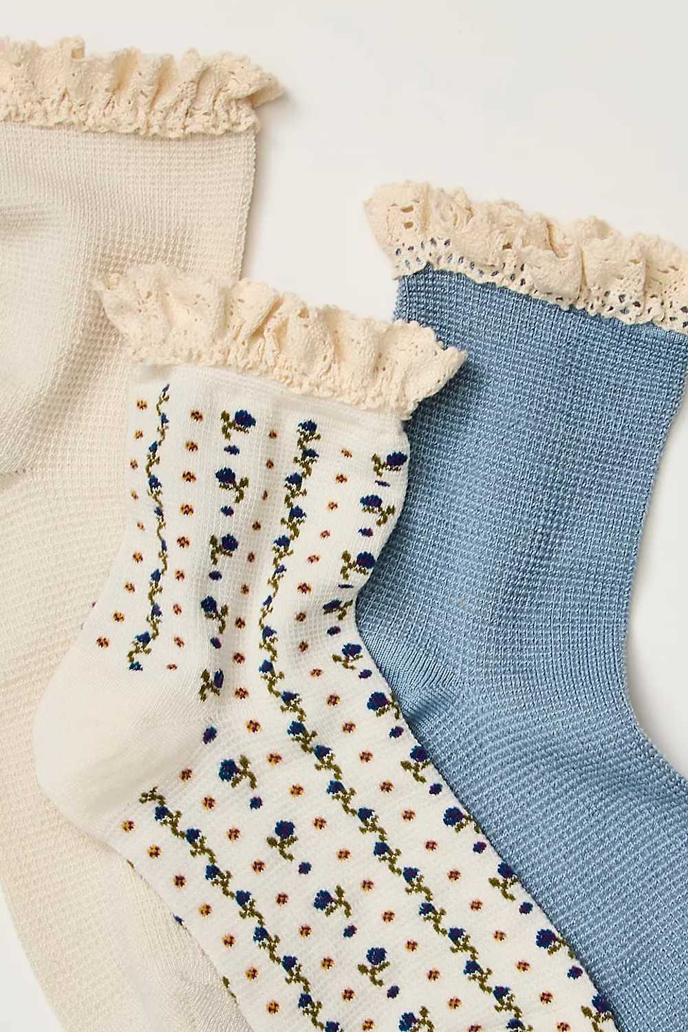 Three pairs of waffle-knit ankle socks with cream ruffled trim, shown overlapping on a light background. One pair is cream with blue and yellow floral embroidery, and another is light blue with small orange polka dots.